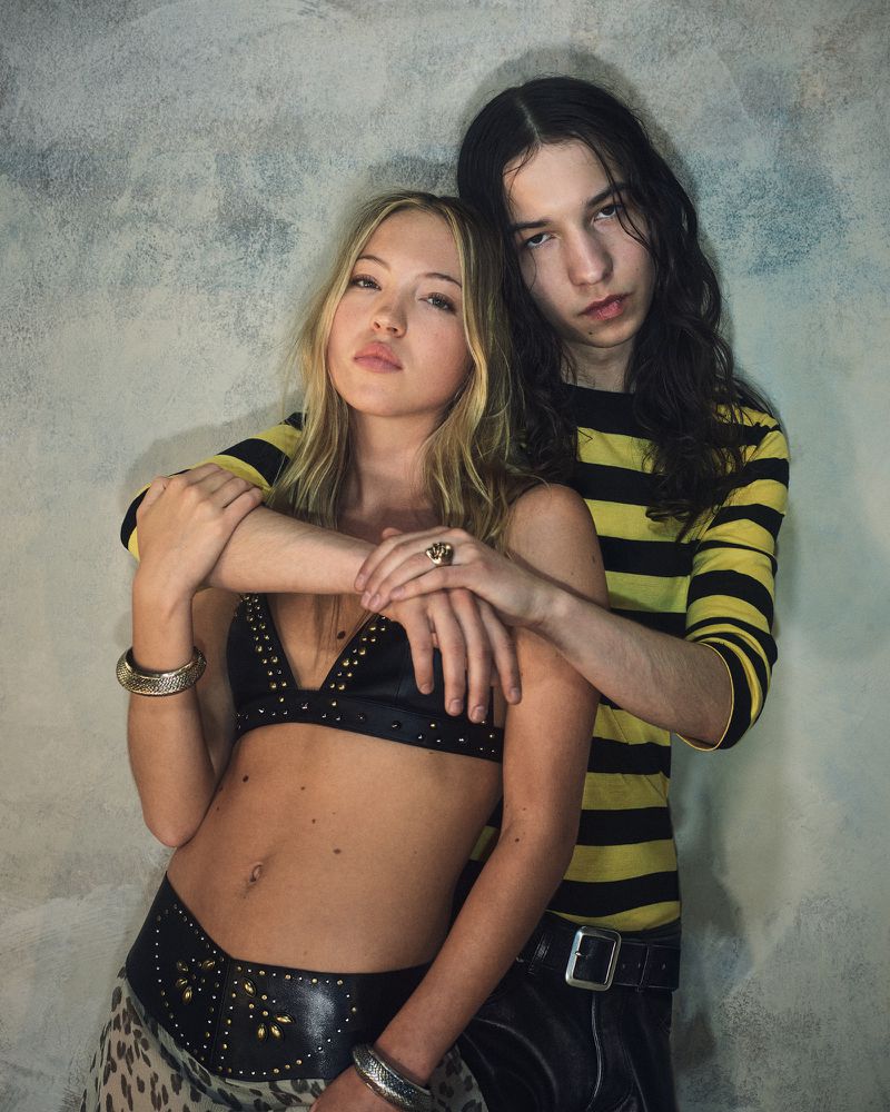 Stylish young couple poses indoors. Woman wears studded bra top and belt; man wears striped yellow and black shirt.