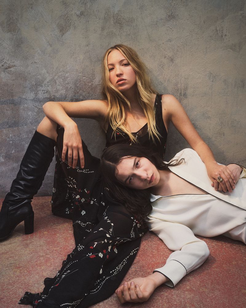 Two women pose together in a studio setting. One woman with blonde hair sits while the other with dark hair leans against her.