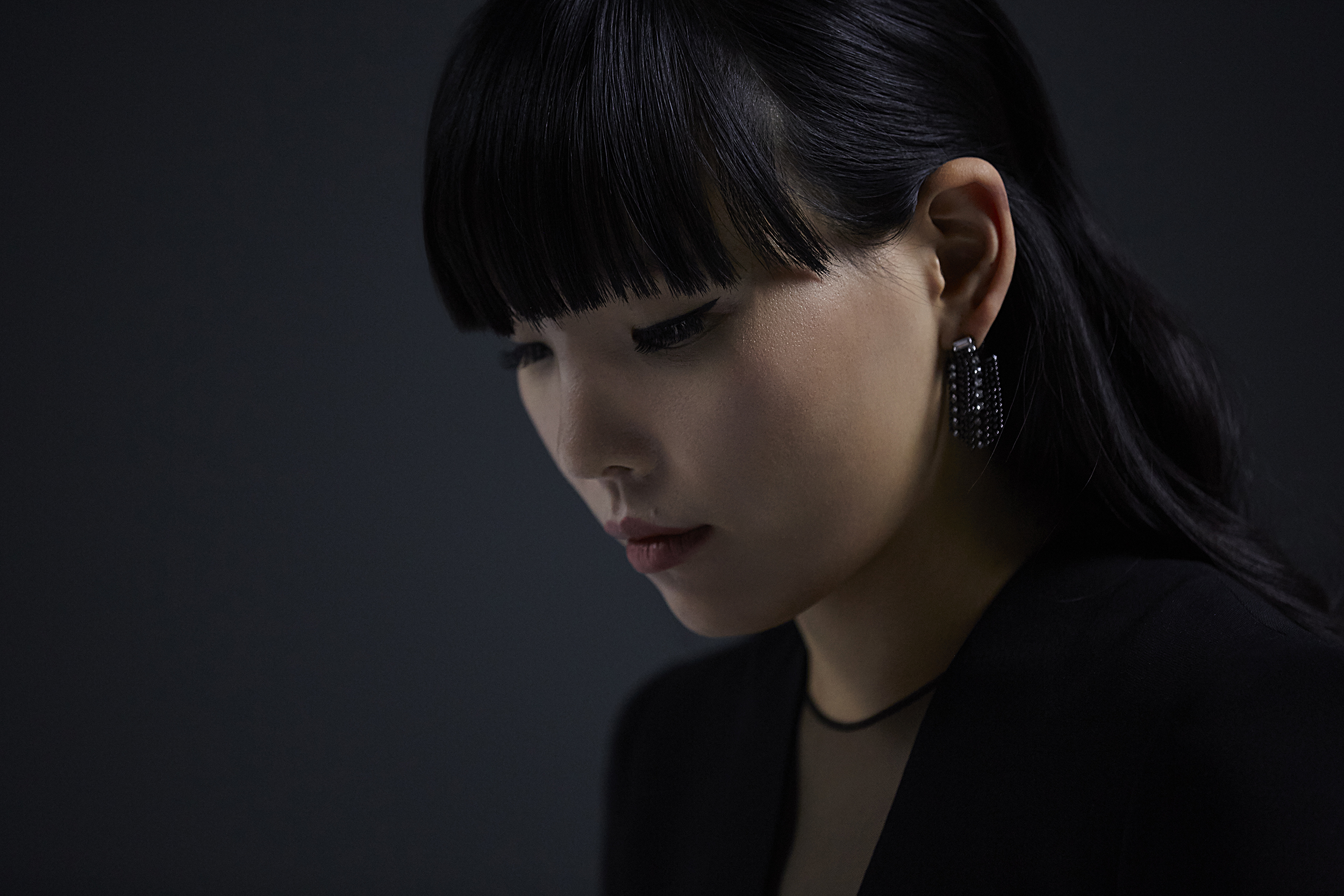 Close-up portrait of a young Asian woman with black hair and dark clothing, featuring a dark background and statement earrings.