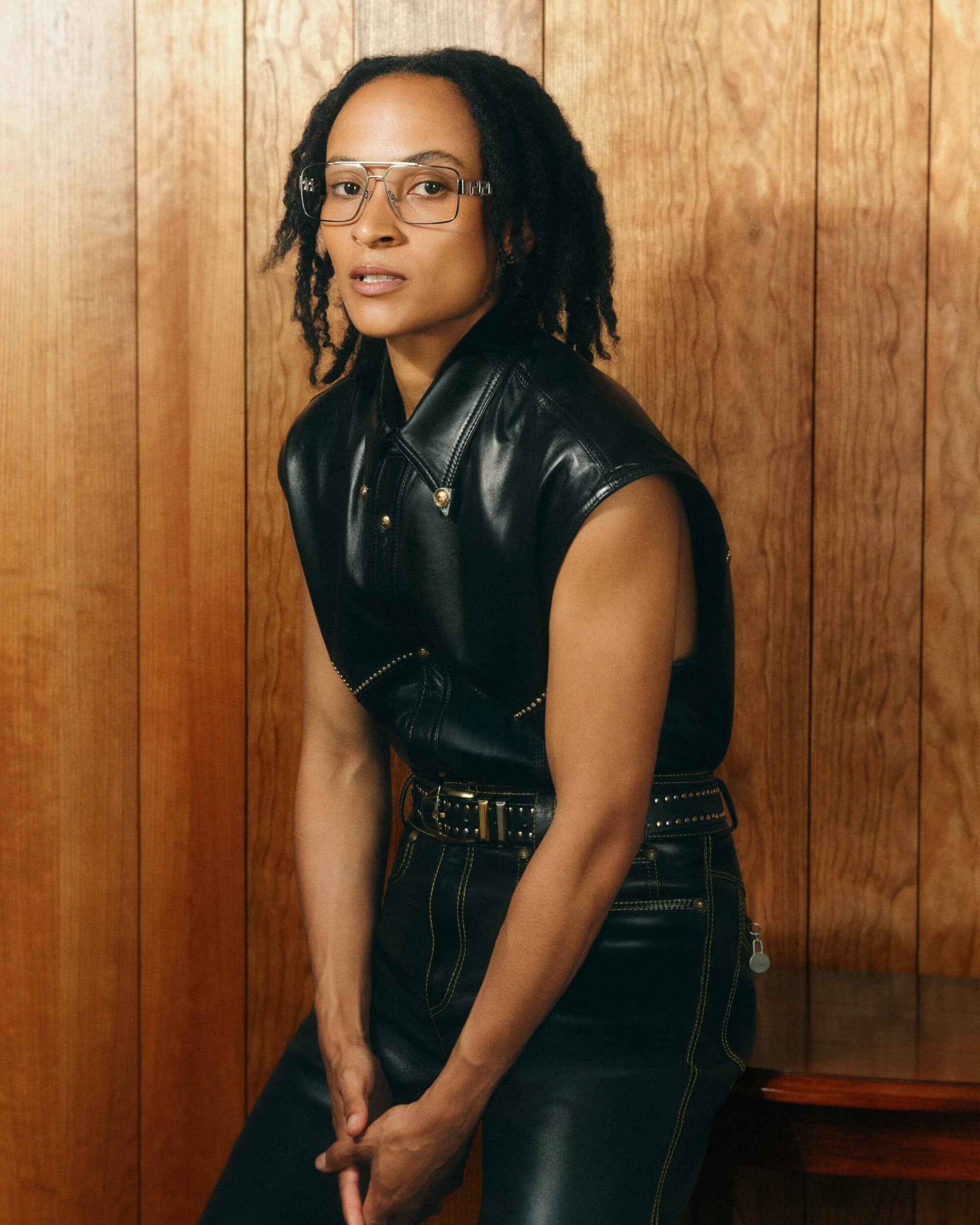 Model wearing a black leather sleeveless shirt and matching trousers with glasses against a wooden panel wall
