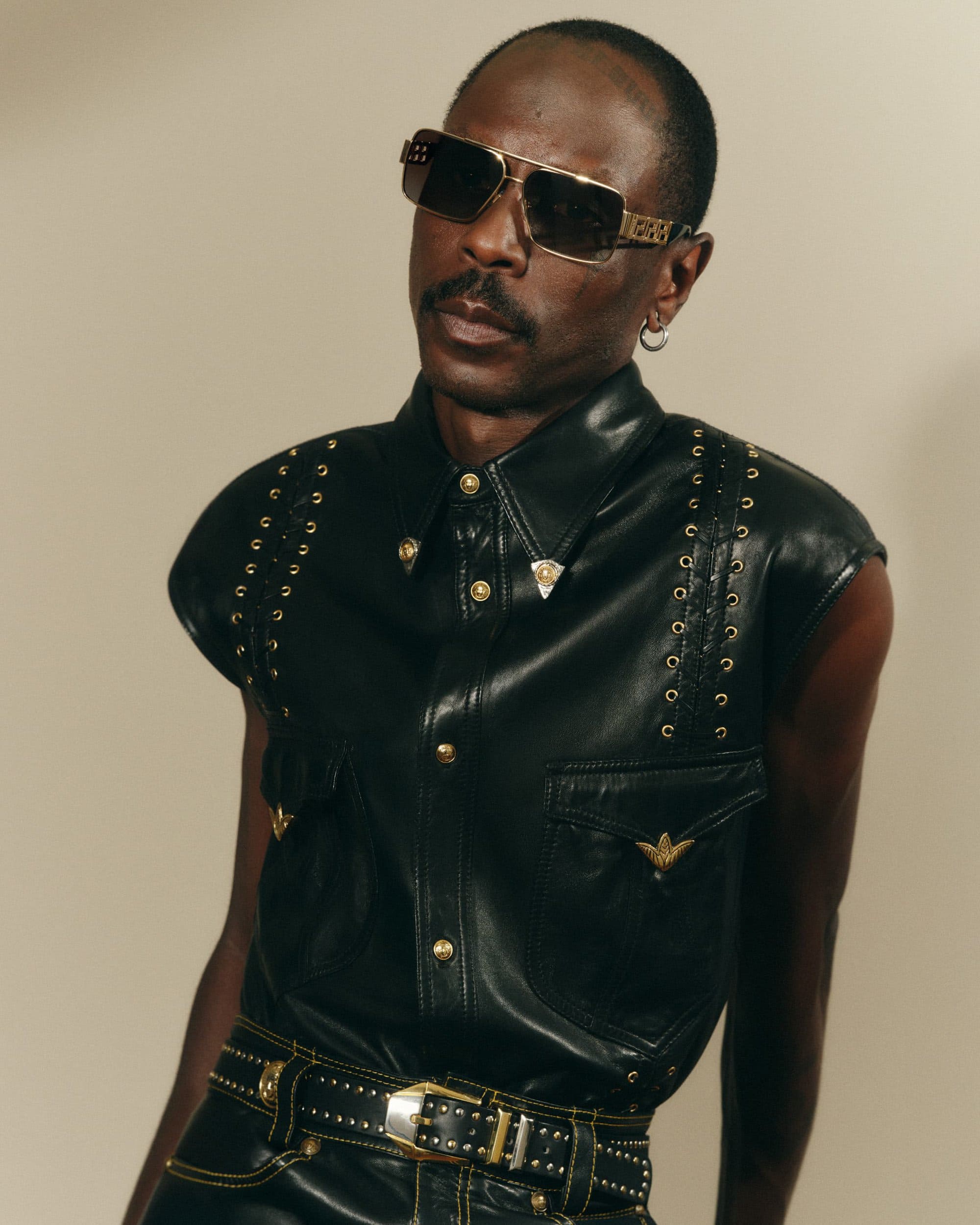 Man wearing sunglasses and a black leather studded shirt against a neutral background for Versace