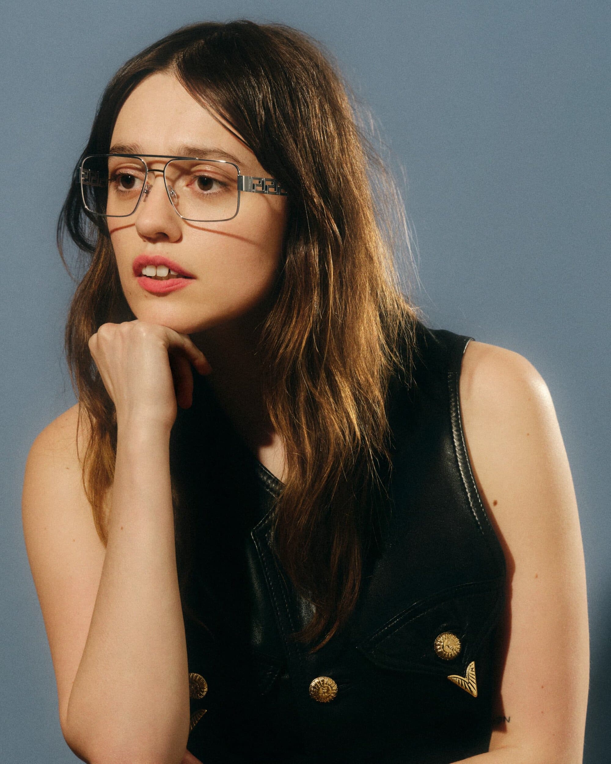 Woman wearing wire-frame glasses and a black leather vest resting her chin on her hand against a grey studio backdrop.