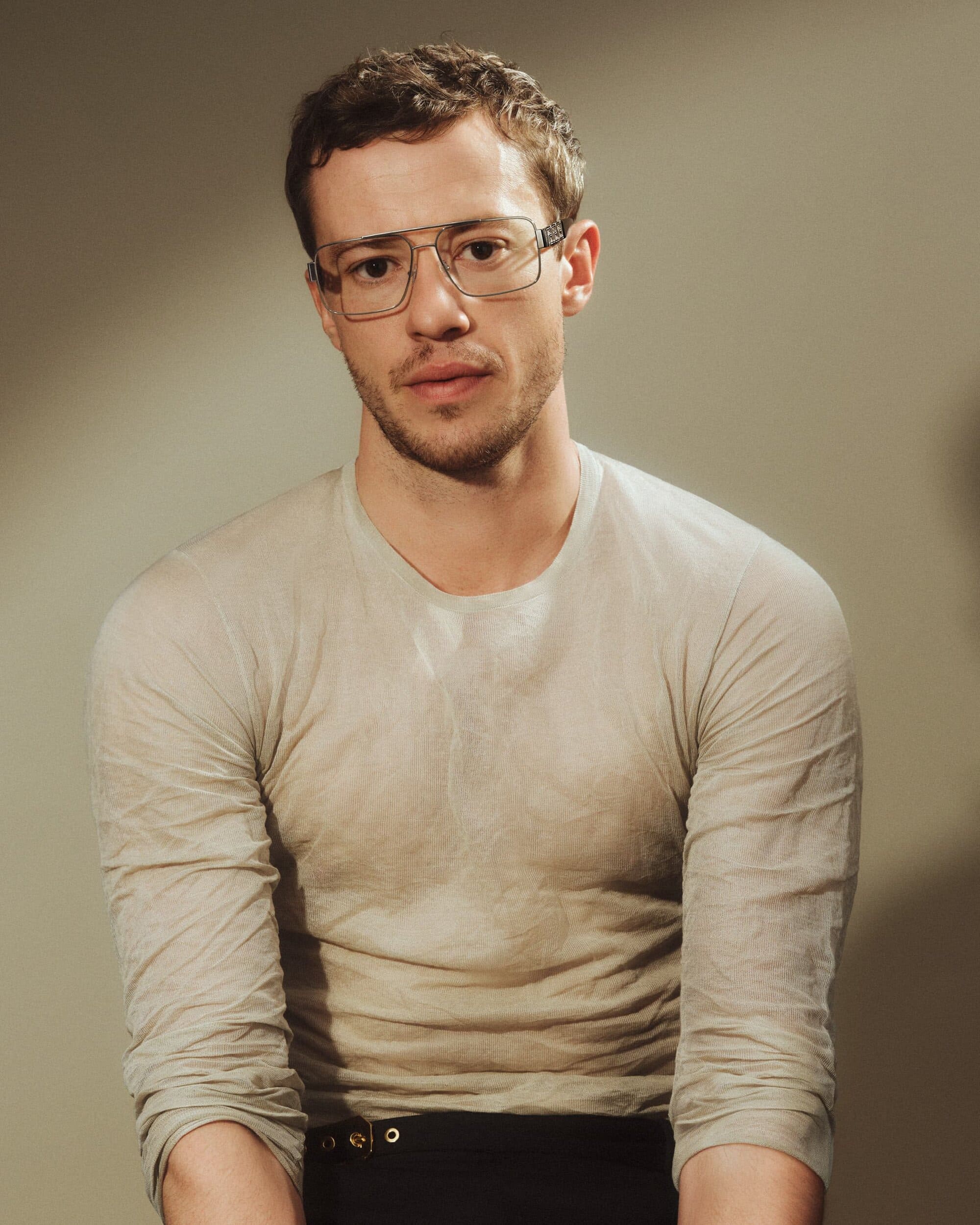 Portrait of Joseph Quinn wearing large square eyeglasses and a fitted light top against a neutral studio background