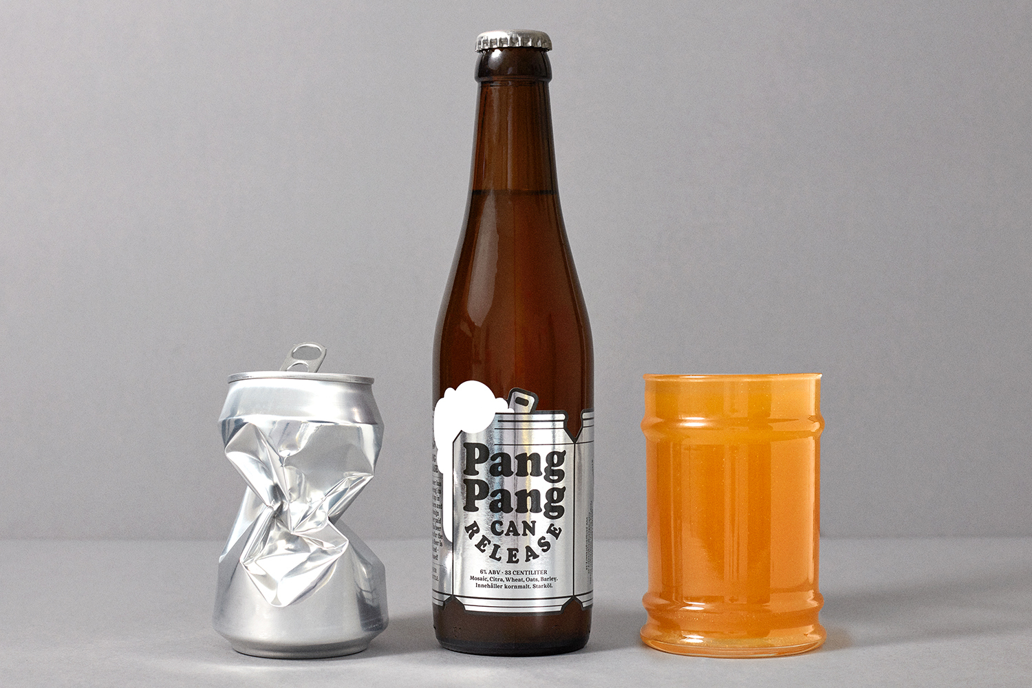Brown beer bottle flanked by a crushed silver can and an amber glass filled with orange liquid against a grey backdrop.