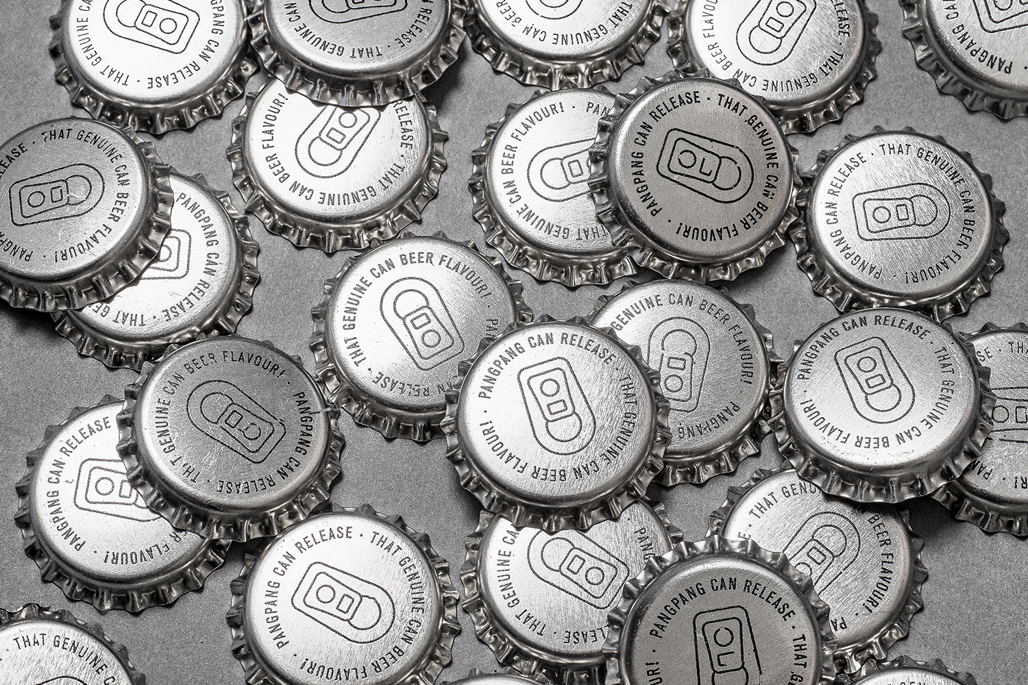 Close-up overhead view of multiple silver metallic PangPang Brewery bottle caps scattered on a solid grey surface.