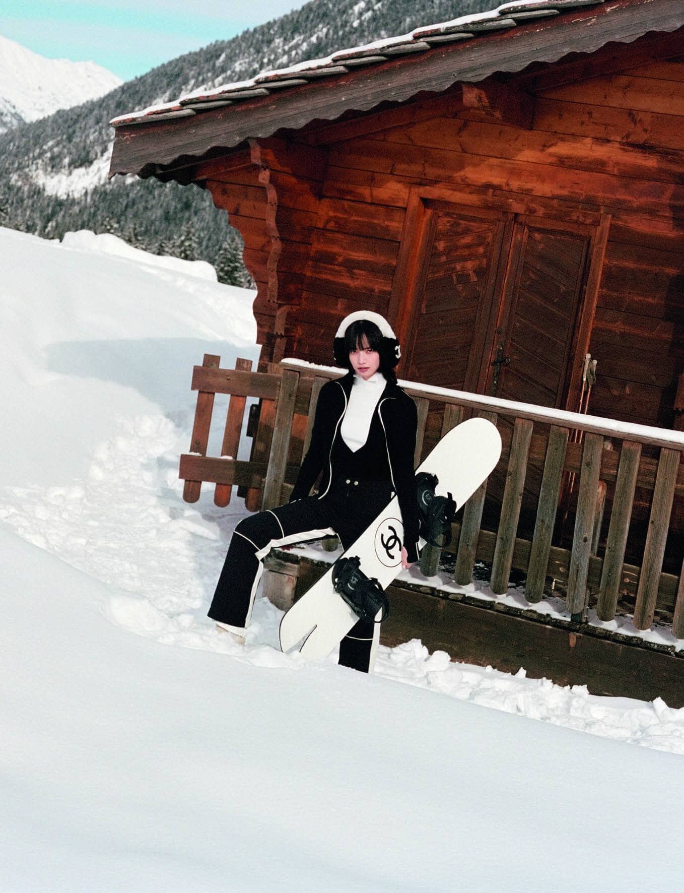 Model Nana Komatsu holding a white snowboard in deep snow outside a wooden alpine chalet for Chanel Coco Neige.