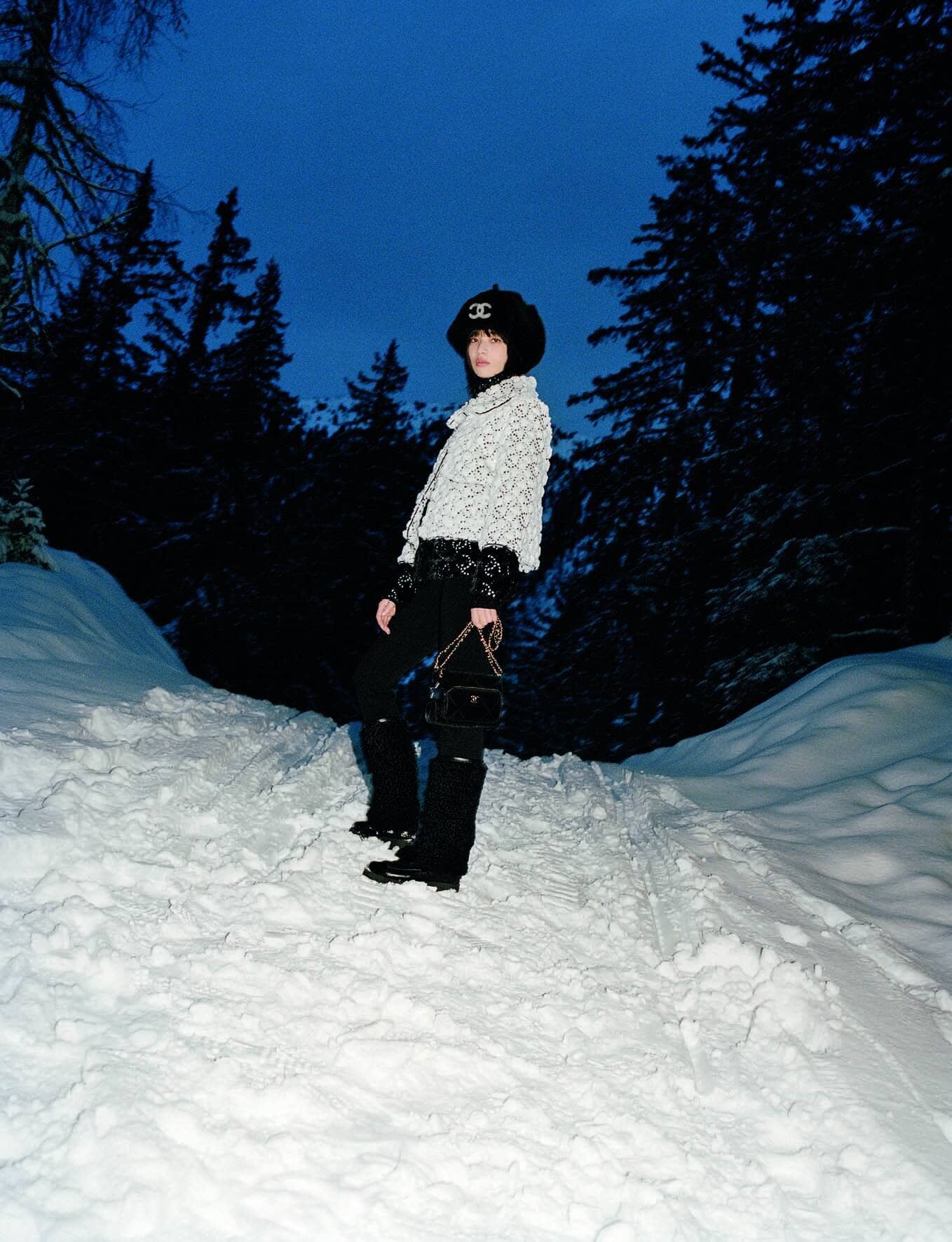 Nana Komatsu wearing Chanel winter fashion standing on a snow-covered path framed by dark evergreen trees at dusk.