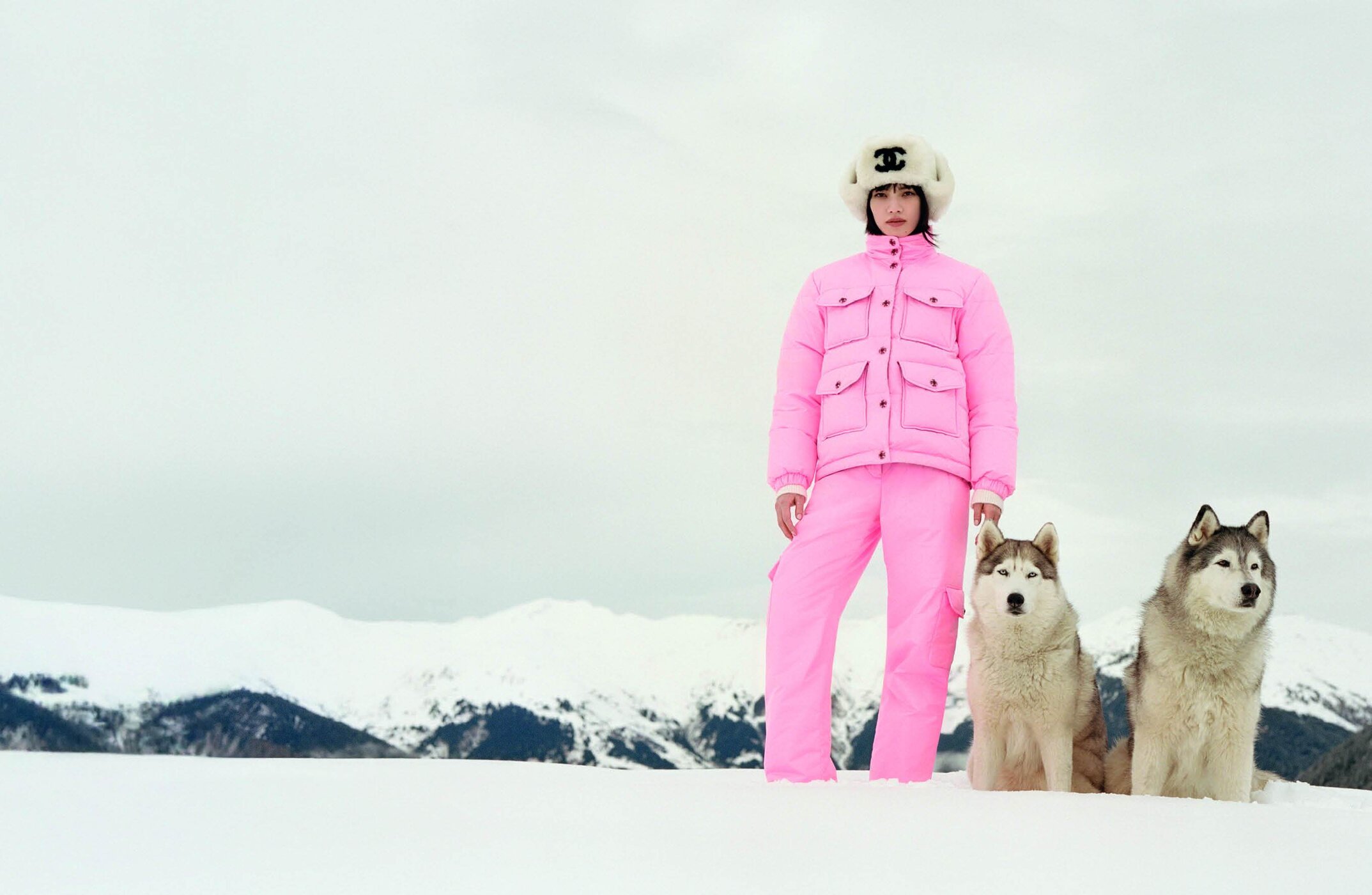 Model in bright pink ski suit and white furry ushanka hat standing with two huskies in snowy mountains