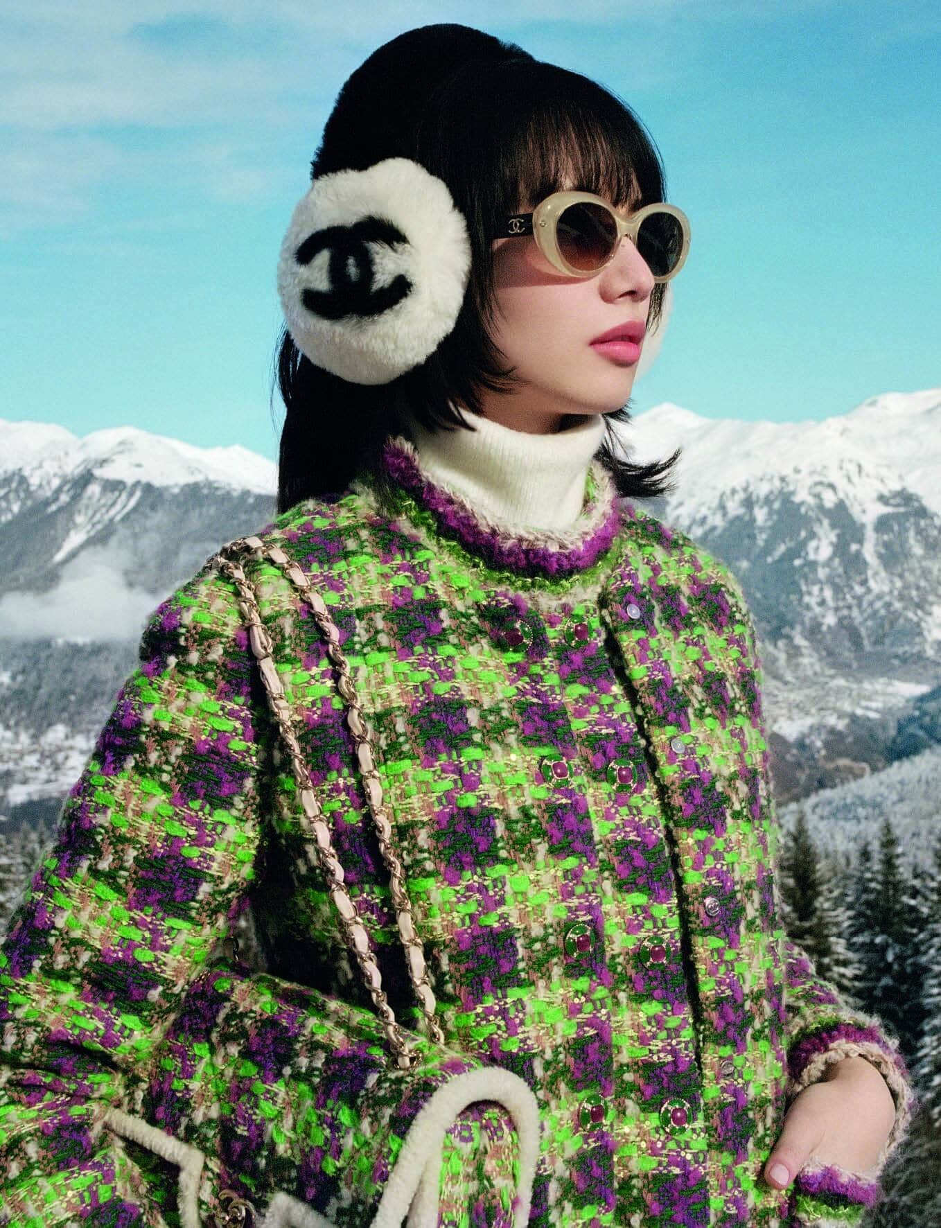 Nana Komatsu wearing a green patterned tweed jacket and white earmuffs with a Chanel logo against a snowy mountain landscape backdrop