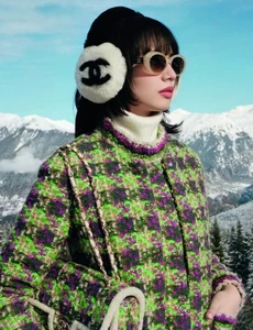 Nana Komatsu wearing a green patterned tweed jacket and white earmuffs with a Chanel logo against a snowy mountain landscape backdrop