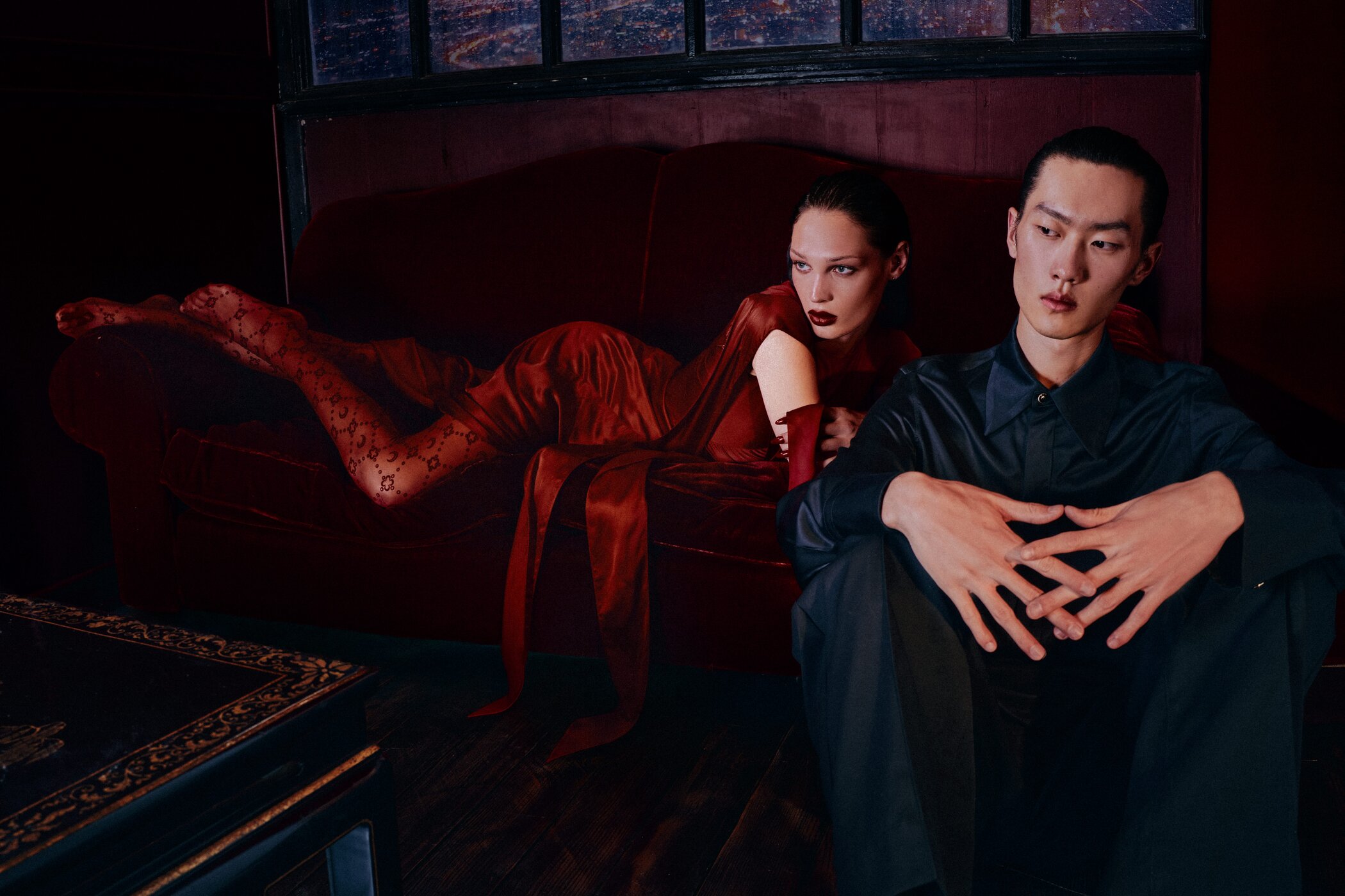 Two models in a dark room: a woman in a red dress reclining on a sofa and a man in a dark shirt seated in the foreground.