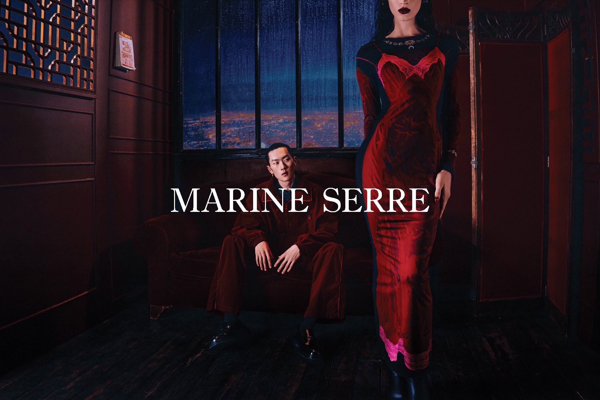 Model in red slip dress standing while a man sits on a velvet couch inside a dark room with a nighttime city window view.
