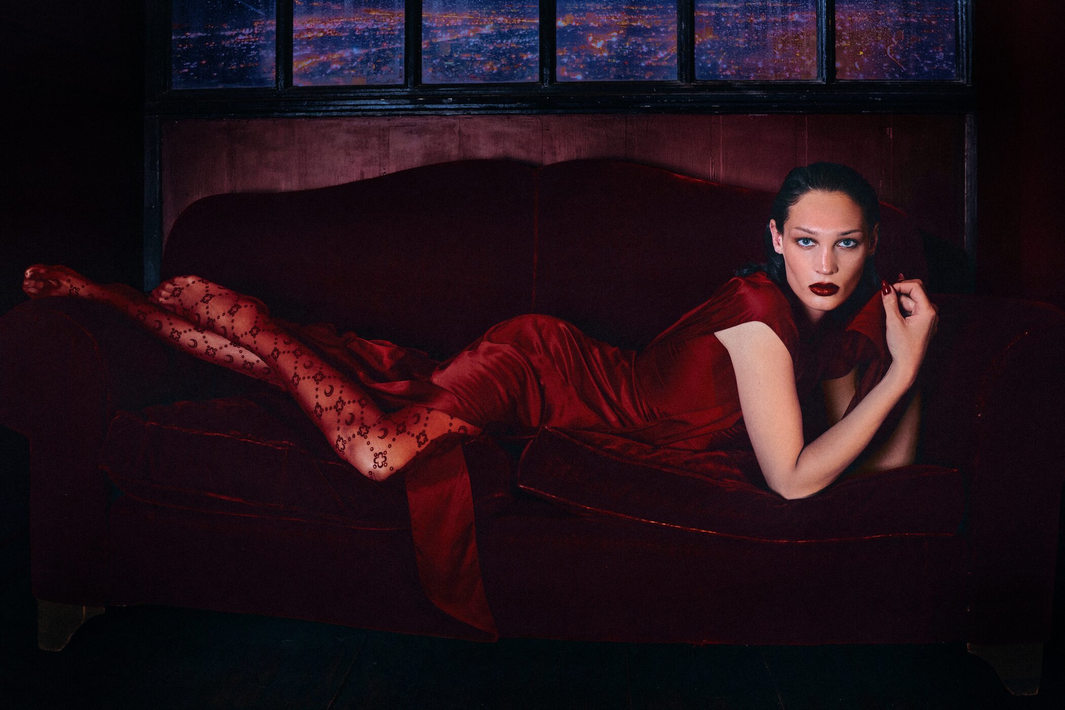 Model in a red dress and patterned tights reclining on a dark red velvet sofa against a moody interior background