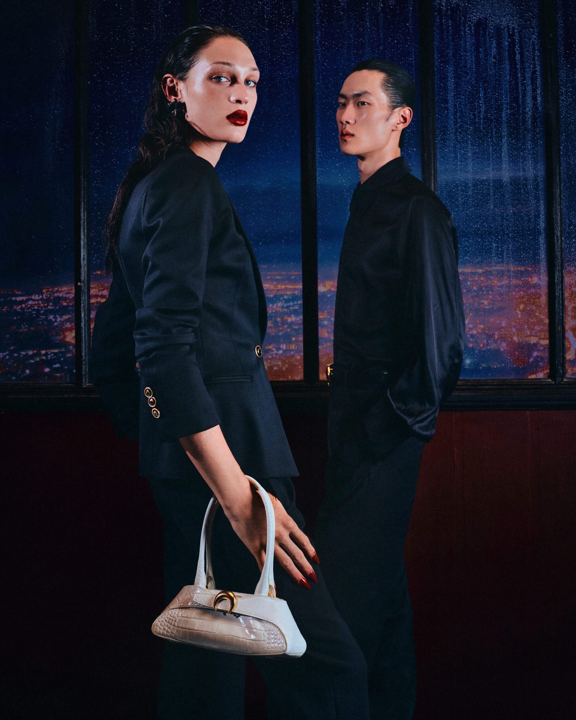 Two models wearing dark attire standing indoors before a large window overlooking a city night scene, holding a structured white handbag.