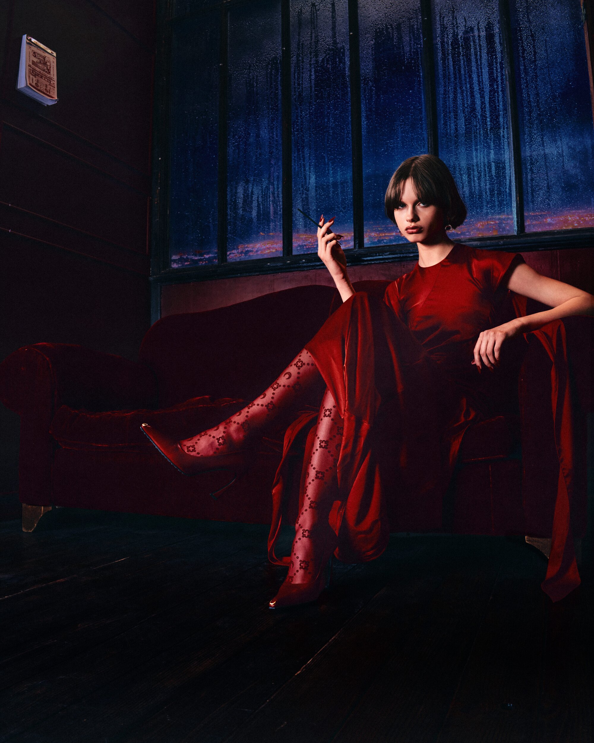 Model in a red dress seated on a red velvet couch in front of a window depicting a rainy city night.