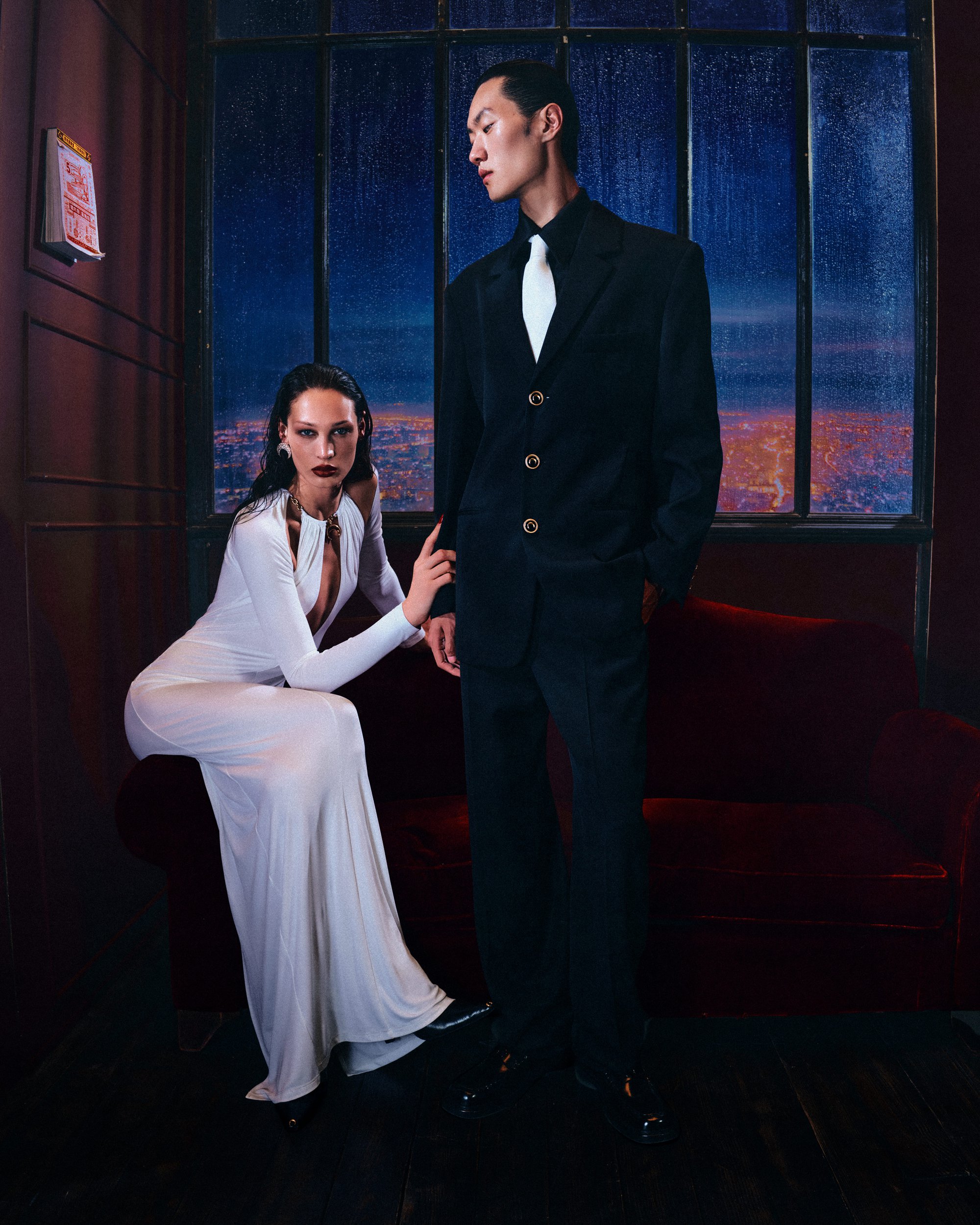Two models posed before a window showing a rainy city view, featuring a seated woman in white and a standing man in black suit.