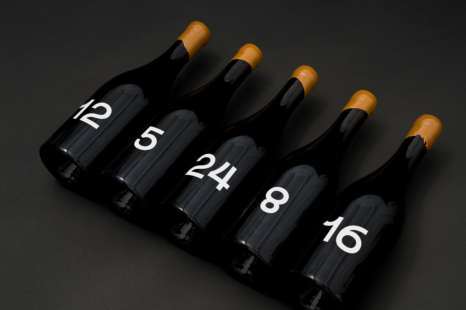 Five dark glass wine bottles with gold wax caps and large white numbers aligned diagonally on a dark grey surface.
