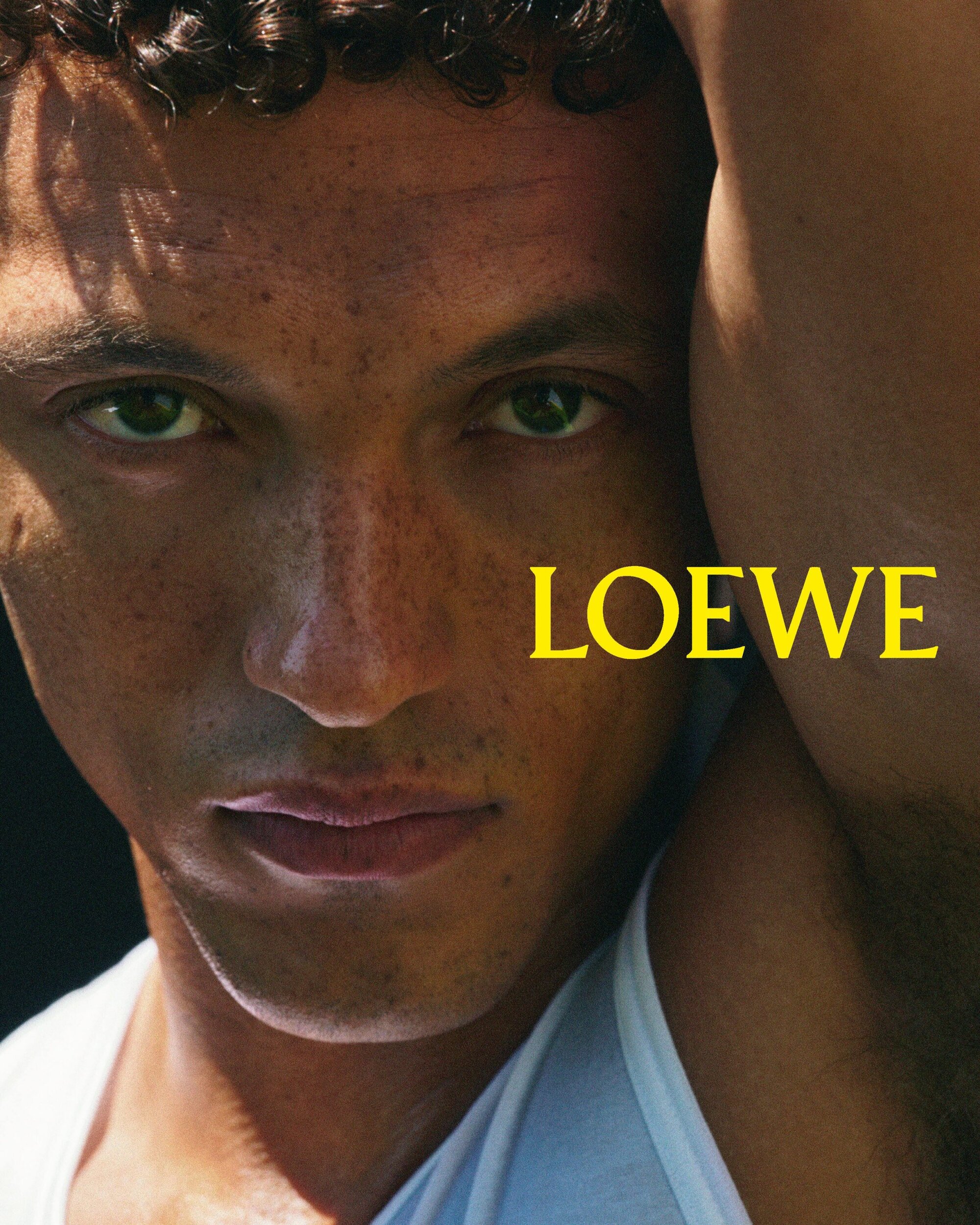 Close-up portrait of a young man with curly hair and freckles wearing a white tank top for Loewe
