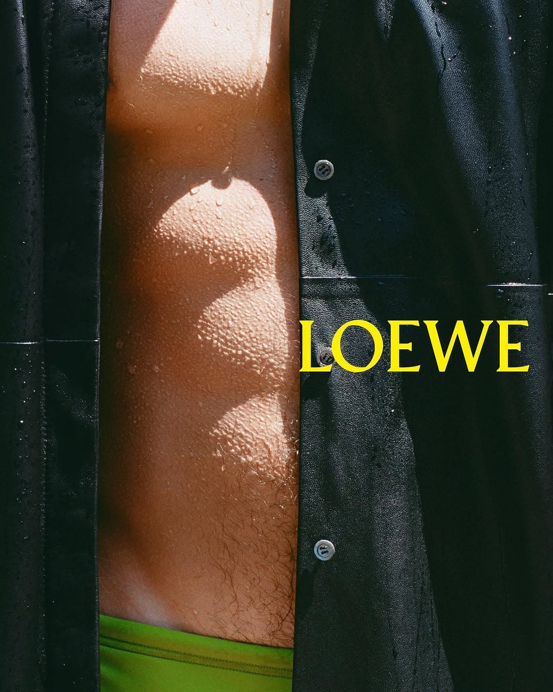 Close-up portrait of a man wearing an open black shirt and bright green swim shorts with water droplets on skin for Loewe.