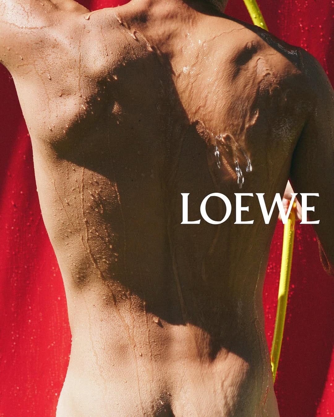 Close-up portrait of a wet person's back against a bright red backdrop with white Loewe text and a yellow pole