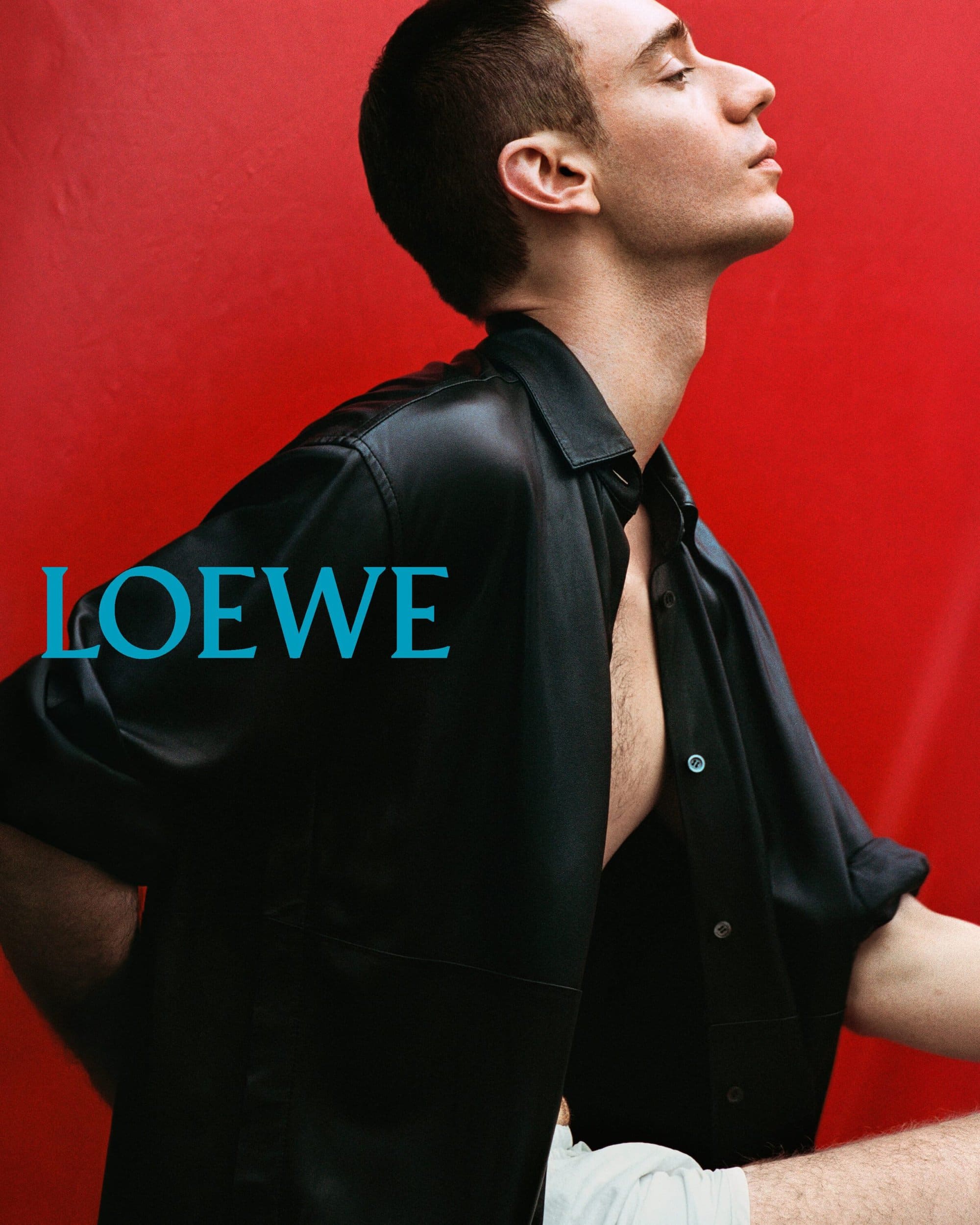 Man in a black leather shirt posing against a bold red background with turquoise Loewe typography