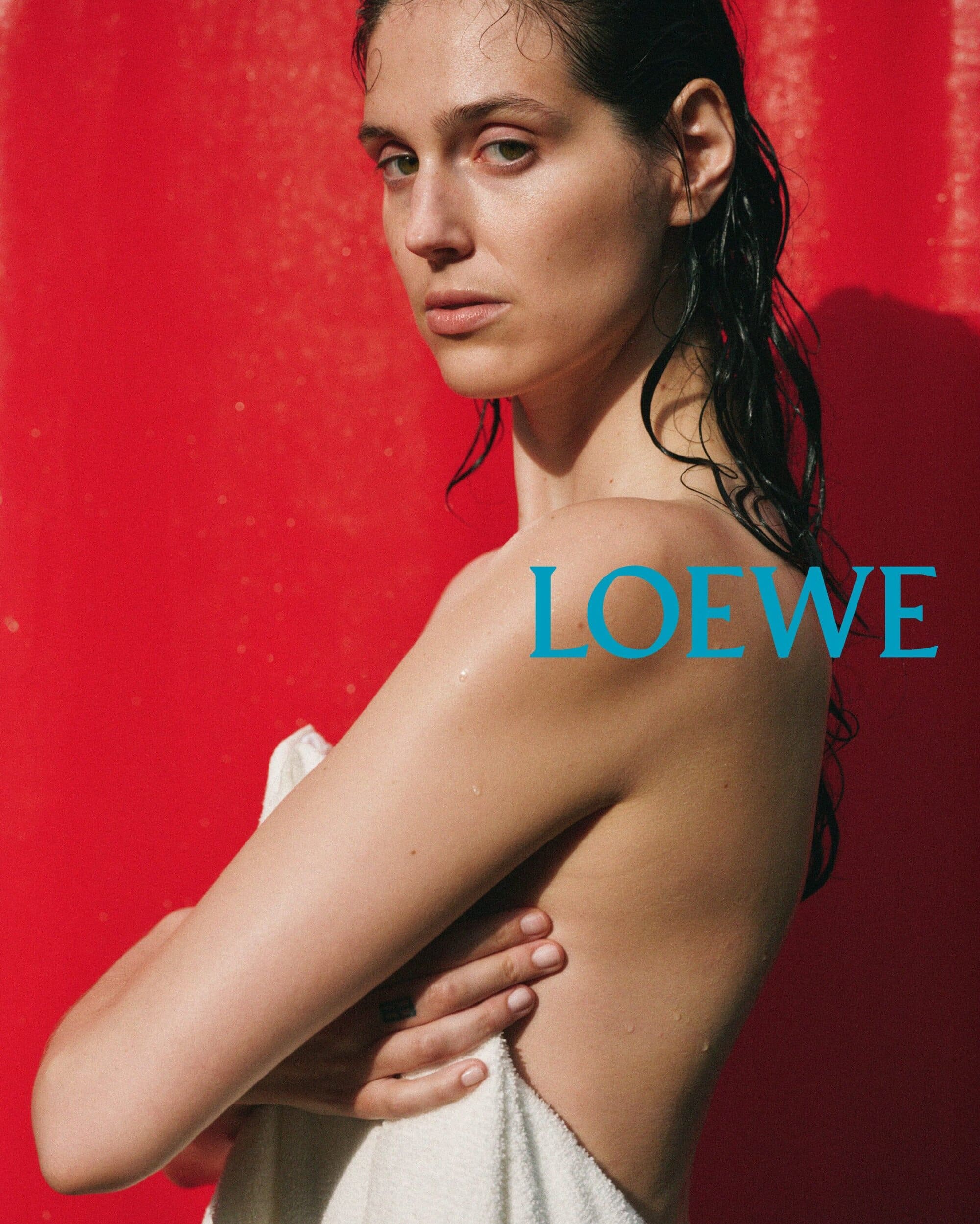 Woman with wet dark hair holding a white towel against a vibrant red background in a Loewe ad campaign.