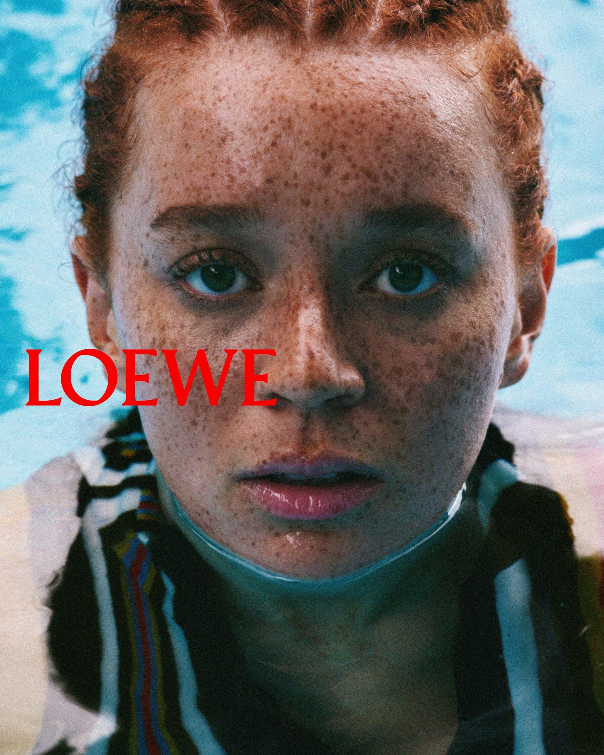 Close-up portrait of a woman with red hair and freckles submerged in blue water with the Loewe logo visible
