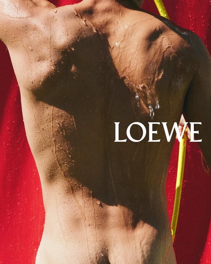 Close-up portrait of a wet person's back against a bright red backdrop with white Loewe text and a yellow pole