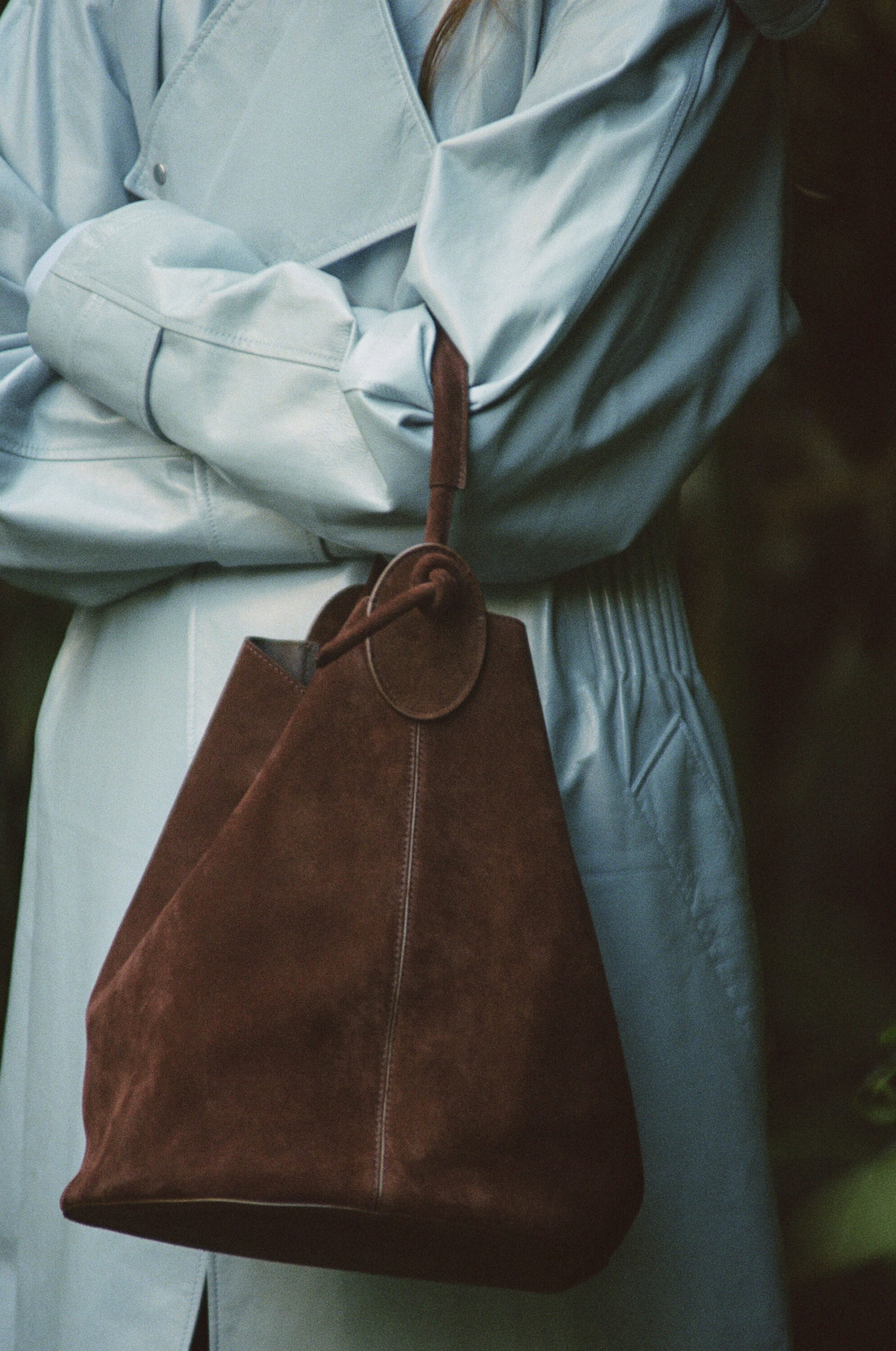 Brown suede bucket bag held against a light blue leather coat in a campaign by Mansur Gavriel