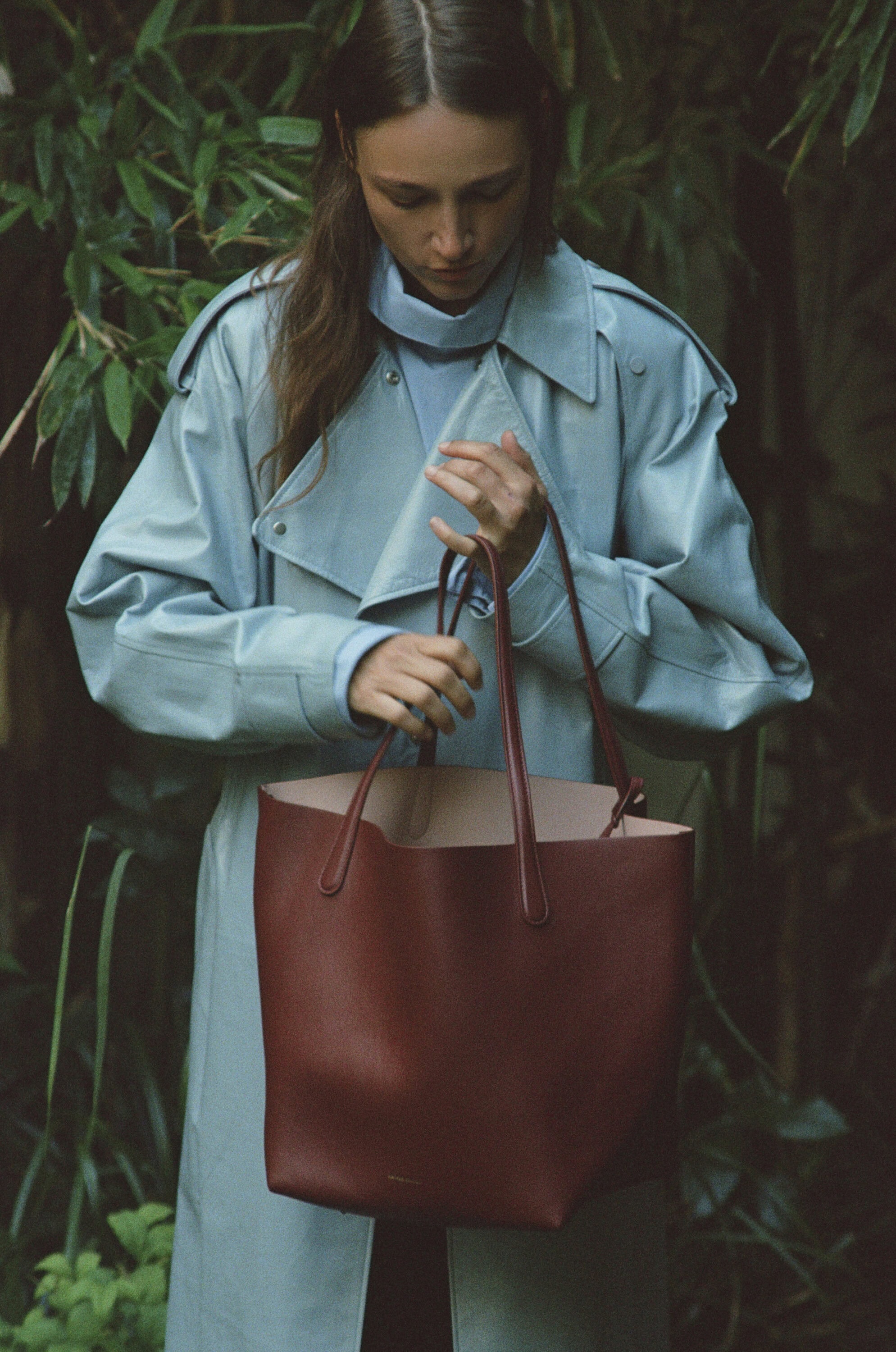 Model wearing a light blue coat holding a large brown leather tote bag amidst green foliage.