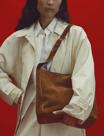 Model wearing a white shirt and cream coat holding a brown suede shoulder bag against a bright red background.