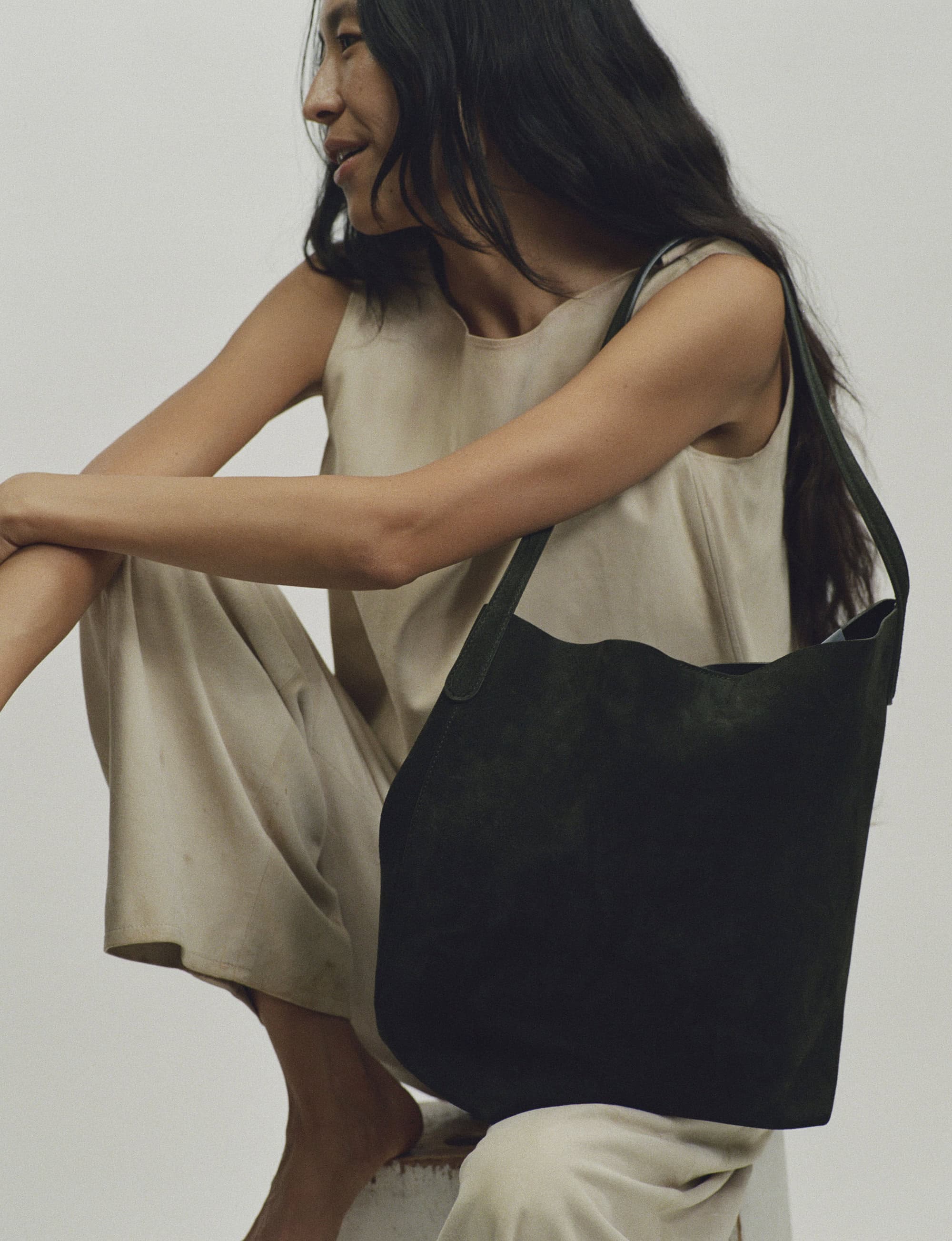 Woman in cream top and trousers holding a dark green leather shoulder bag against a neutral background