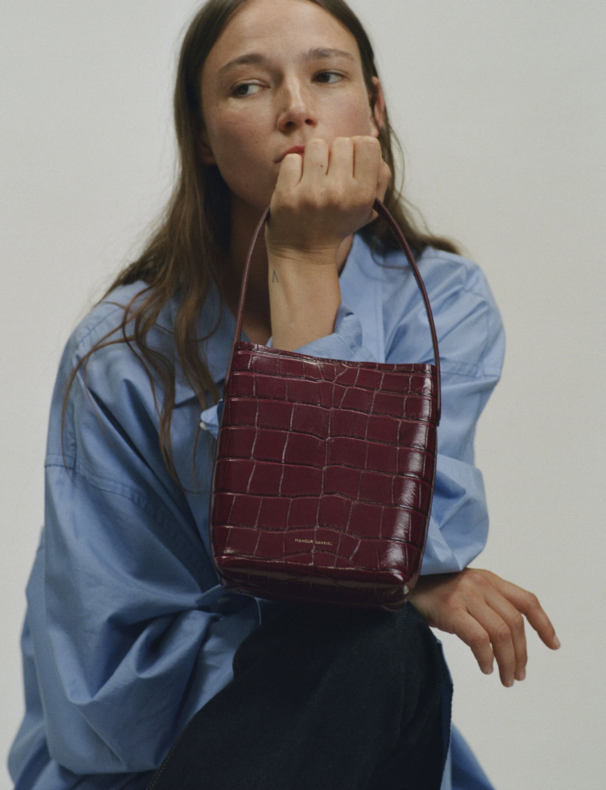 Woman wearing a blue button-down shirt holding a glossy burgundy embossed leather handbag against a neutral grey background