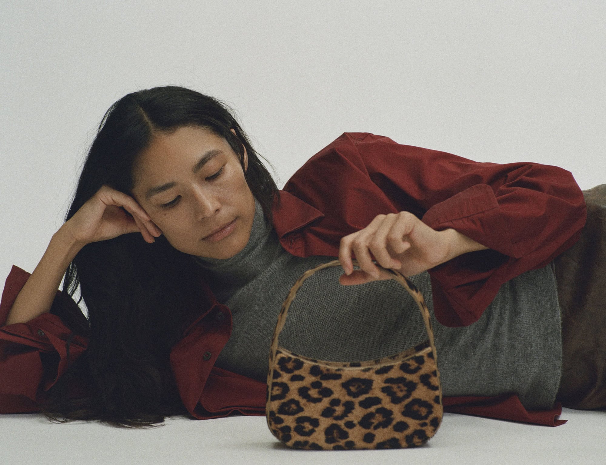 Woman in a grey turtleneck and red overshirt reclining on a light background while holding a leopard print handbag