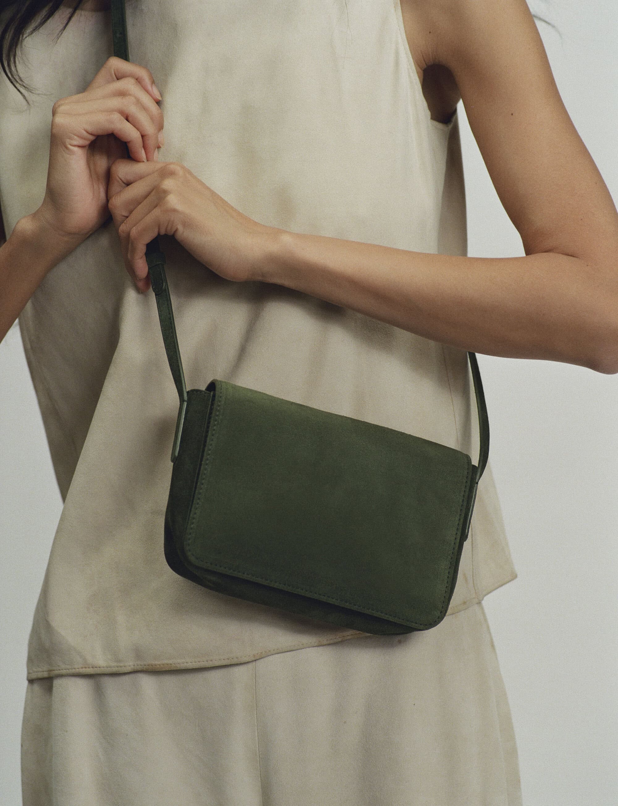 Model wearing a neutral sleeveless top holding the strap of a dark green leather crossbody bag against a plain background