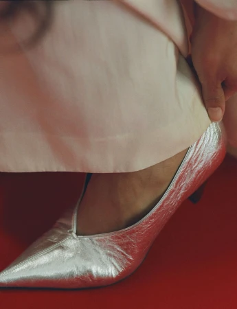Close-up of a person wearing silver metallic pointed-toe heels and pale trousers against a solid red background for Mansur Gavriel.