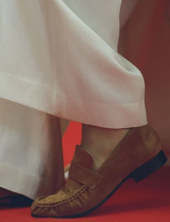 Close-up of a brown suede loafer worn with white trousers against a warm red background.