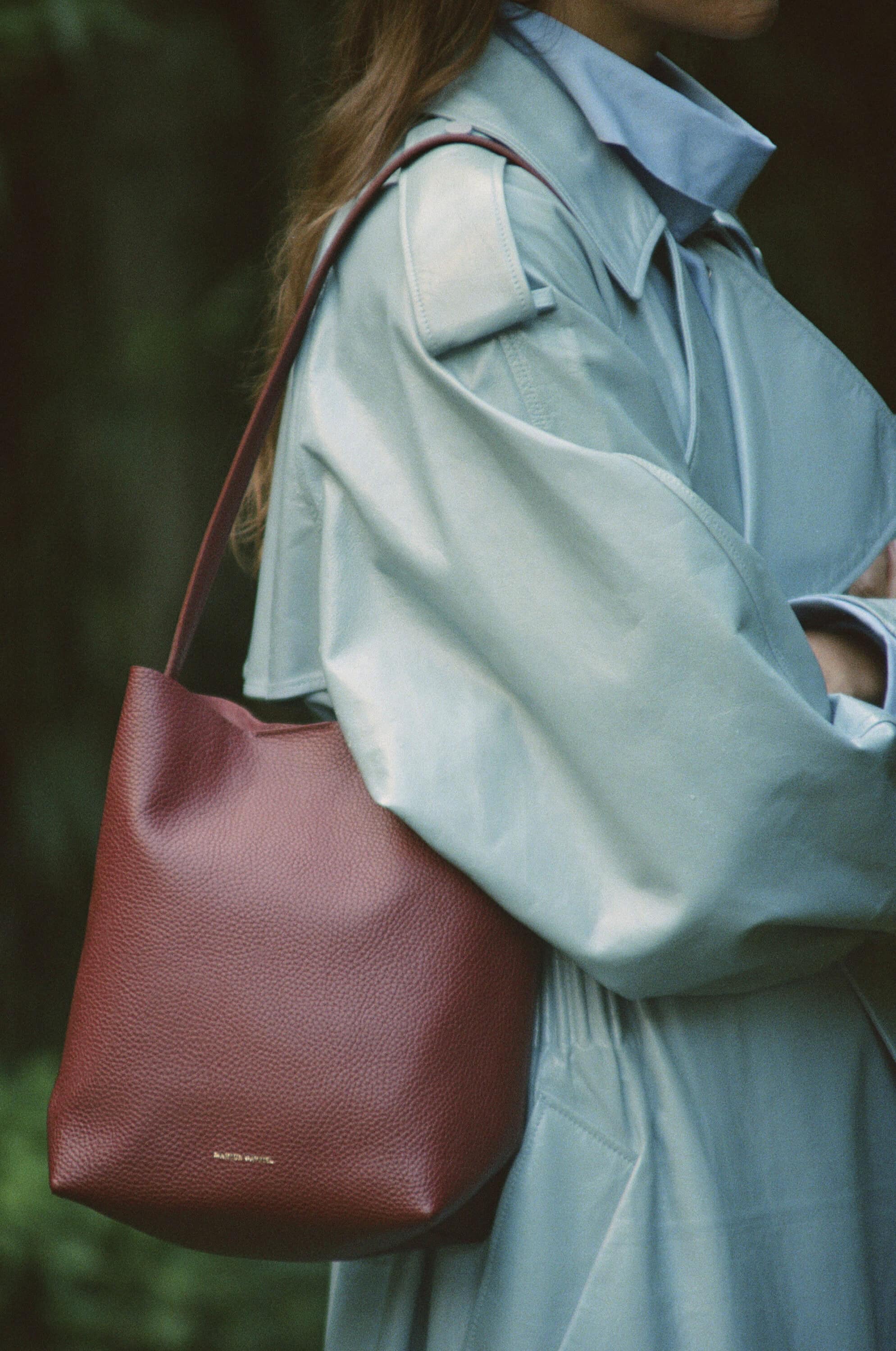 Model wearing a light blue coat and carrying a burgundy leather shoulder bag outdoors