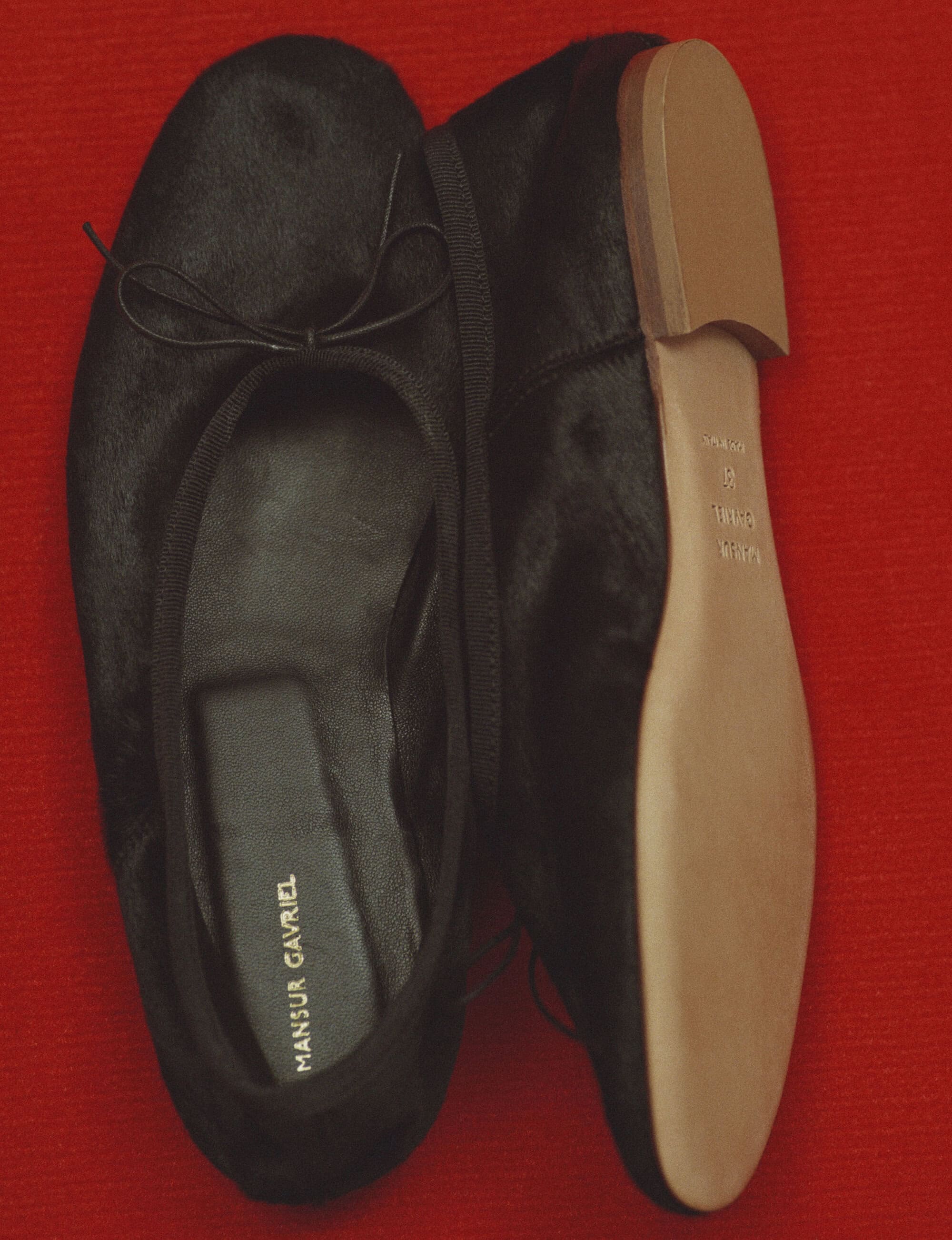 Top-down view of a pair of black ballet flats with tan soles resting on a textured red surface for Mansur Gavriel