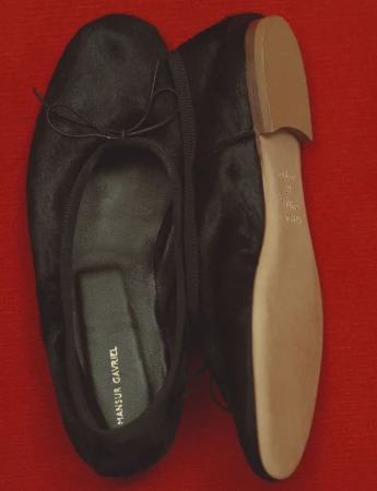 Top-down view of a pair of black ballet flats with tan soles resting on a textured red surface for Mansur Gavriel