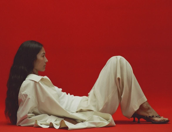 Model in cream trousers and matching jacket reclining against a solid vibrant red background.