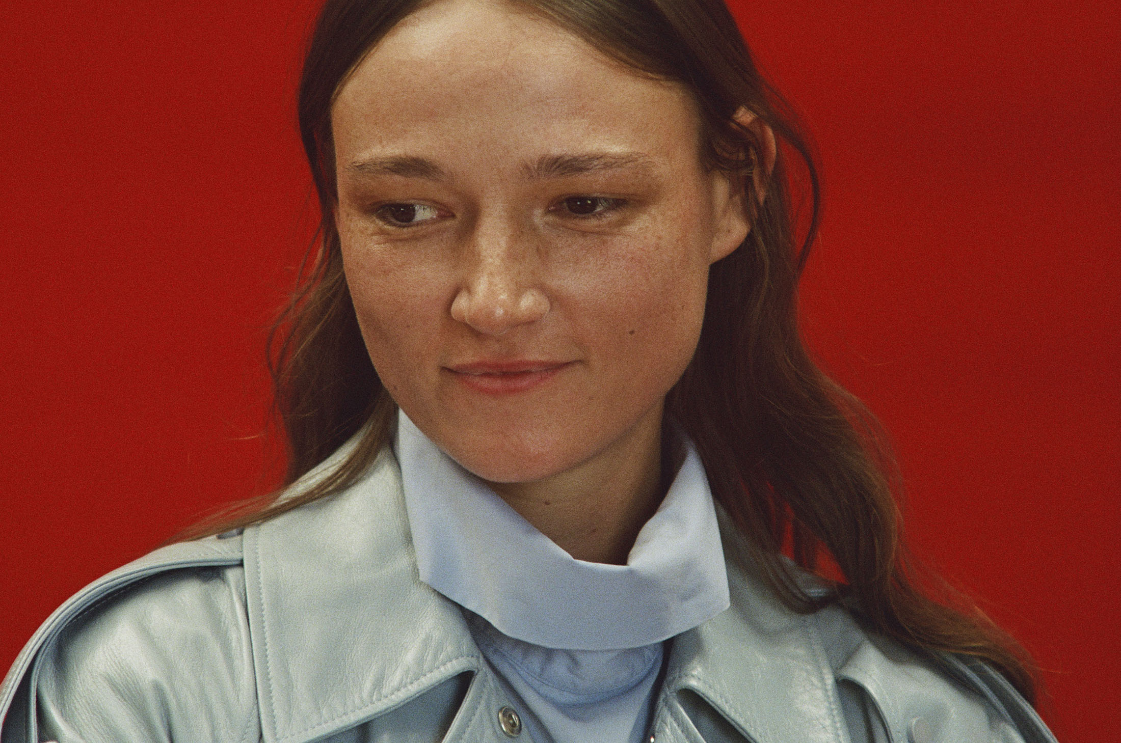 Close-up portrait of a woman wearing a light blue leather jacket against a vibrant red backdrop for Mansur Gavriel Fall 2025.