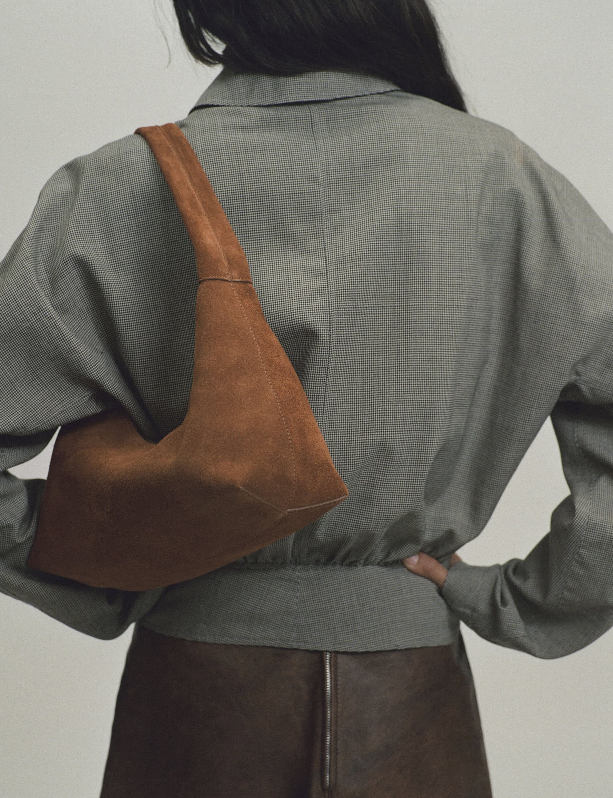 Rear view of a person wearing a grey shirt carrying a brown suede shoulder bag against a plain background