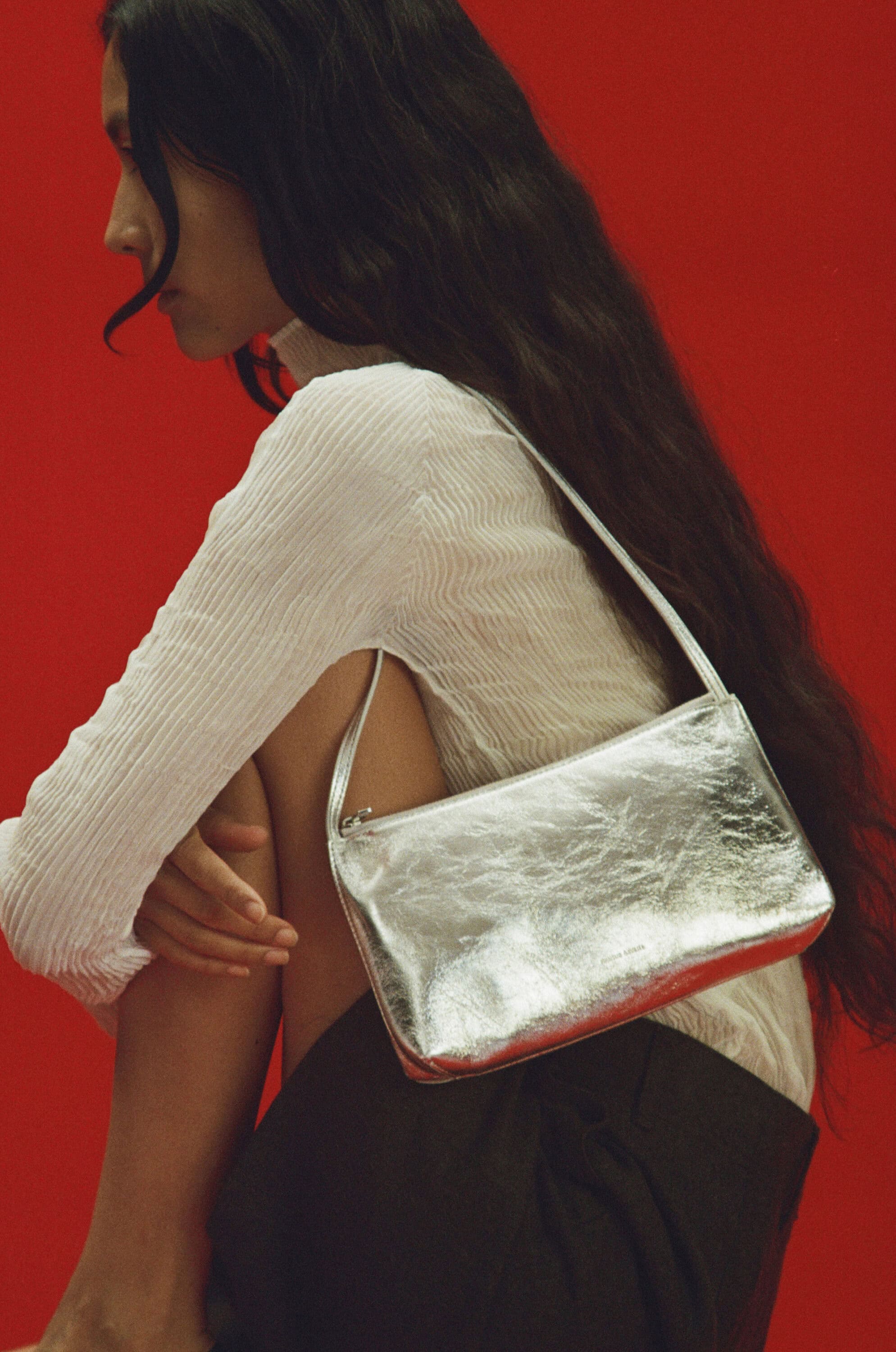 Model with long dark hair wearing a white textured top holding a metallic silver shoulder bag against a vibrant red background.