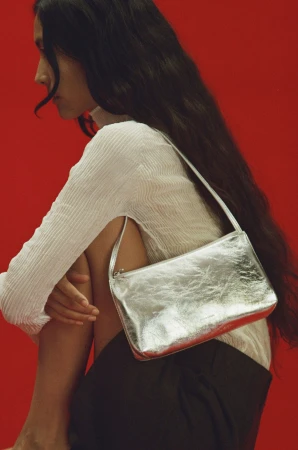 Model with long dark hair wearing a white textured top holding a metallic silver shoulder bag against a vibrant red background.