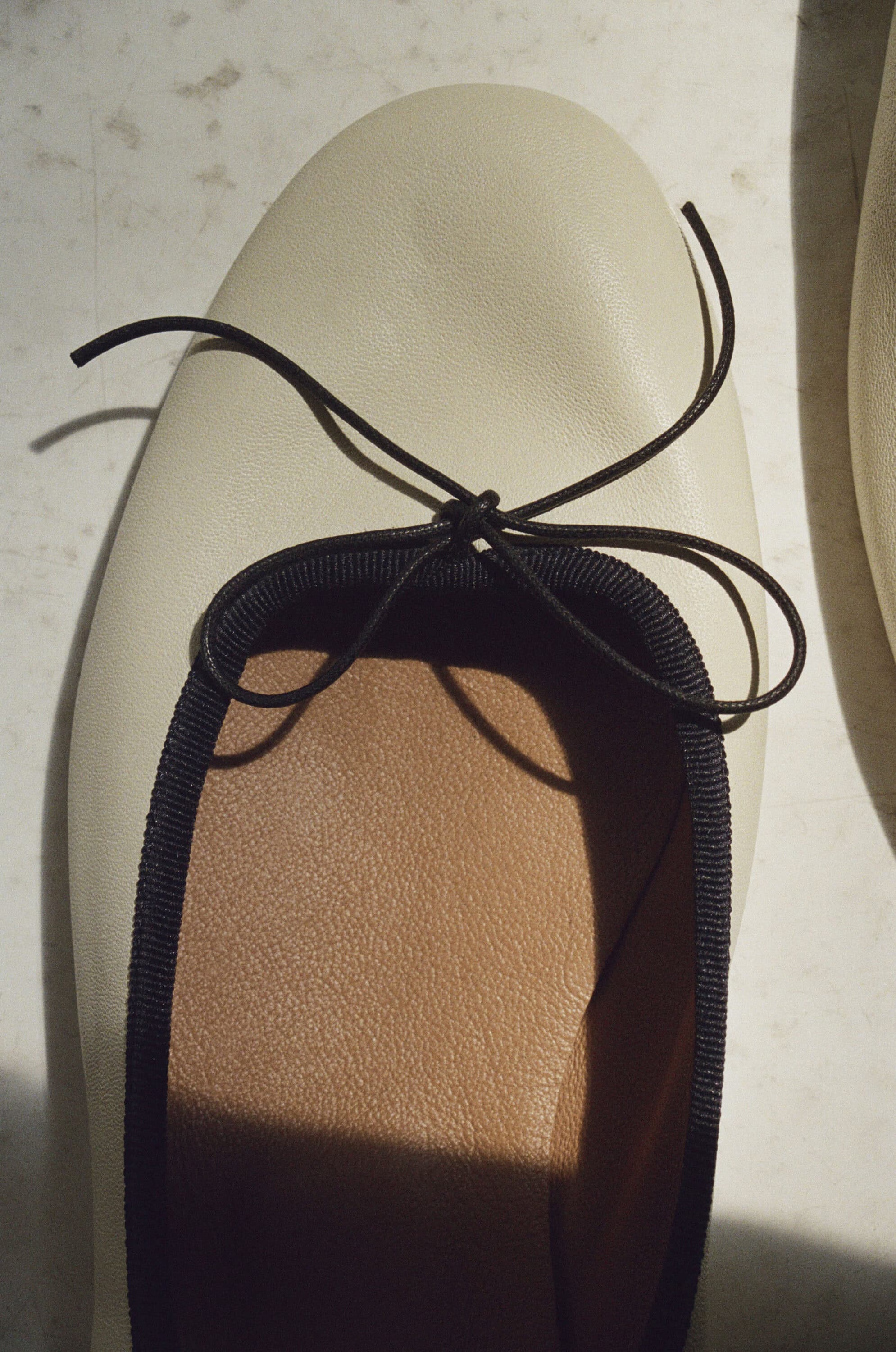 Close-up of a cream-colored leather ballet flat with a thin dark bow and ribbed trim resting on a surface