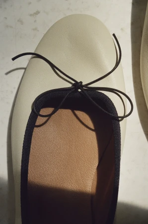 Close-up of a cream-colored leather ballet flat with a thin dark bow and ribbed trim resting on a surface