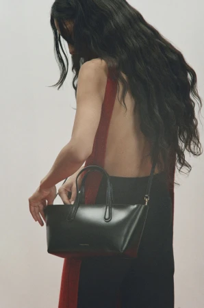 Model with long dark wavy hair holding a black leather handbag against a neutral background for Mansur Gavriel Fall 2025 ad campaign.