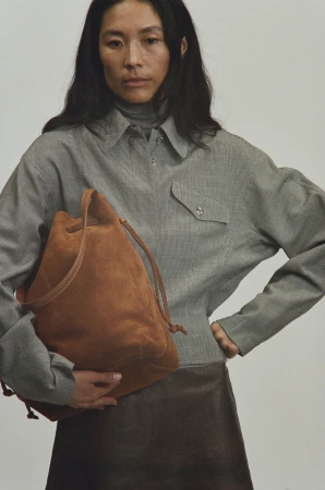 Woman holding a brown suede bucket bag wearing a grey patterned shirt against a plain studio background.