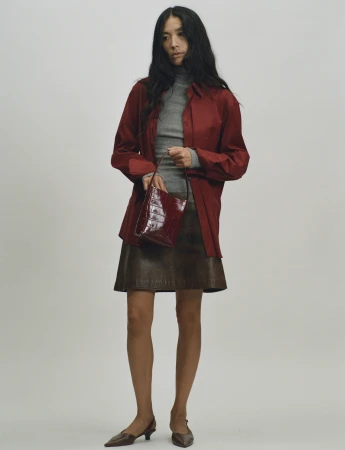 A woman holding a burgundy leather handbag wearing a red shirt, grey turtleneck, brown skirt, and dark shoes against a plain white wall